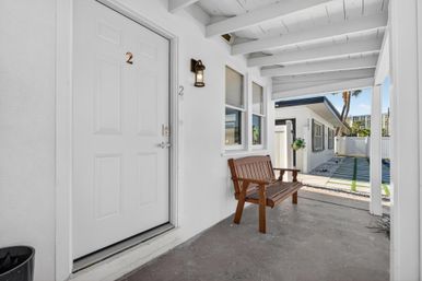Covered white front porch of a coastal cottage unit with door numbered 2, wooden bench, wall lantern, and a walkway lined with gravel and palm trees