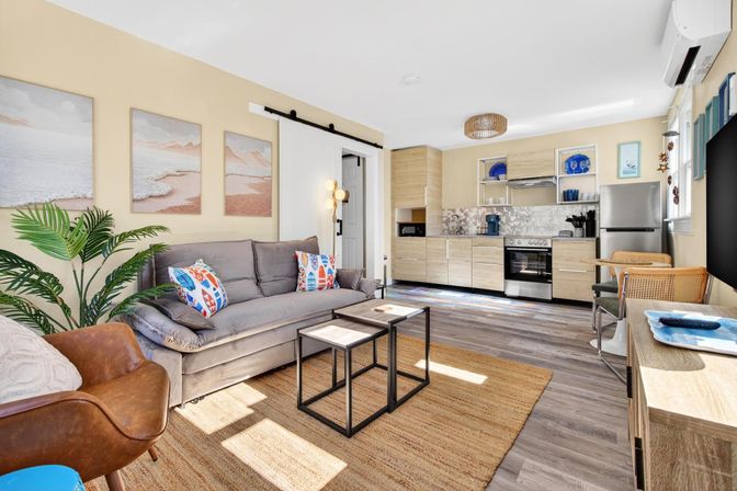Sunlit modern coastal-style studio apartment with open-plan living room and kitchenette, gray sofa with colorful pillows, leather accent chair, potted palm, nested coffee tables, jute rug, and light wood kitchen with stainless steel appliances.