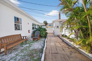 Sunny tropical courtyard with a paver walkway between white buildings, wooden bench and Adirondack chair, palm plants and blue sky