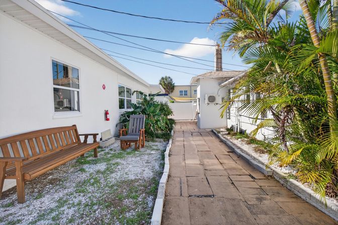 Sunny tropical courtyard with a paver walkway between white buildings, wooden bench and Adirondack chair, palm plants and blue sky