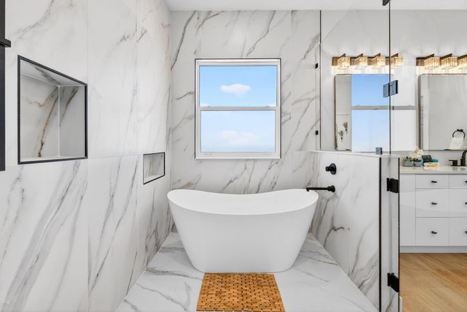 Spa-like modern bathroom with white marble walls and floor, freestanding soaking tub beneath a window with blue sky, matte black fixtures, recessed shower niches, glass shower door and woven wooden bath mat.
