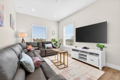 Sunlit modern living room with a large gray sectional sofa topped with pastel pillows and rolled throws, wall-mounted flat-screen TV above a white media console, gold nesting coffee tables on a patterned area rug, potted plant and floor lamp by two bright windows.