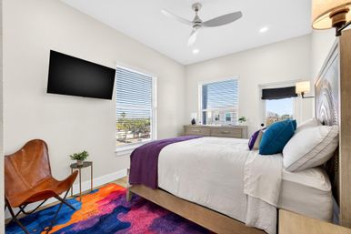 Bright modern bedroom with white linens, purple throw and teal pillows, wall-mounted TV, leather accent chair and vibrant multicolor rug, sunlit windows revealing palm trees outside.