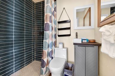 Small modern bathroom with dark blue horizontal subway-tile shower, circular-pattern shower curtain, hexagon tile floor, white toilet, hanging black shelves, gray vanity with mirror and neatly folded towels