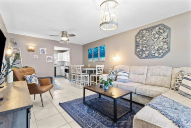 Bright coastal-style open-concept living room and dining area with beige sectional sofa, leather accent chair, blue rug and pillows, wooden coffee table, ceiling fan and pendant light, tile floor, and kitchen visible through doorway.