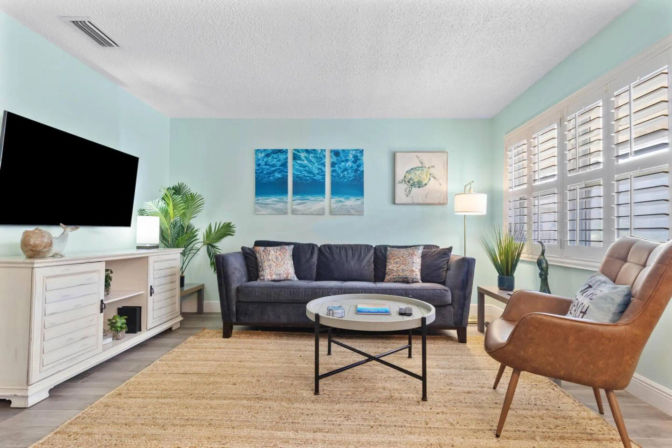 Bright beachy living room with light teal walls, navy sofa, round coffee table on a natural jute rug, tan leather accent chair, white media console with wall-mounted TV, shuttered windows, coastal artwork and indoor plants.