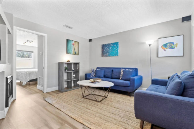 Cozy modern living room with matching blue sofas, round coffee table on a natural jute rug, hardwood floors, colorful wall art, floor lamp and open doorway to a bedroom