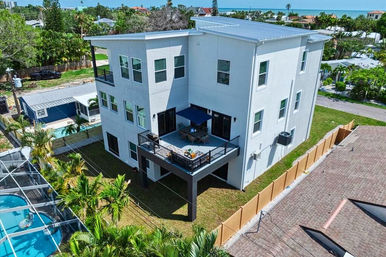 Modern three-story coastal house with raised corner deck featuring an outdoor dining set and blue umbrella, fenced yard with palm trees, a screened neighbor pool, and ocean horizon in the background.