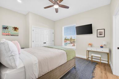 Bright modern coastal-style bedroom with a comfy bed, wooden ceiling fan, wall-mounted TV, white double-closet doors, small desk and window showing neighborhood rooftops.
