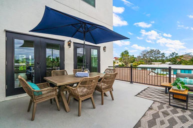 Modern rooftop patio with wooden dining table, six wicker chairs, large navy umbrella, cushioned bench and outdoor rugs overlooking nearby homes under a bright blue sky