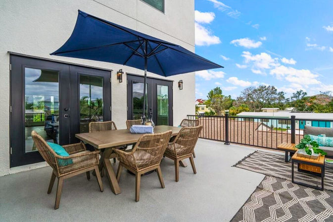 Modern rooftop patio with wooden dining table, six wicker chairs, large navy umbrella, cushioned bench and outdoor rugs overlooking nearby homes under a bright blue sky