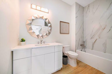 Bright modern white bathroom interior with sleek vanity and undermount sink, geometric round mirror, toilet, bathtub with marble-look tile surround and wood-look flooring