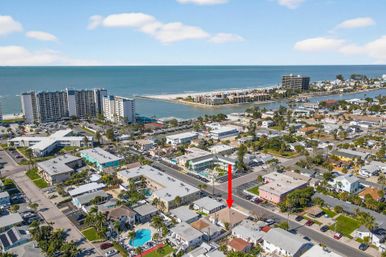 Aerial view of a sunny coastal neighborhood with turquoise ocean, sandy beach and a canal; high-rise beachfront condos in the background and a red arrow pointing to a single-family property two blocks from the shore.