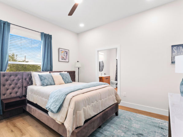 Bright modern bedroom with tufted upholstered bed, layered cream and teal bedding, teal curtains at a large window, ceiling fan, hardwood floors, patterned rug and an open doorway leading to an ensuite bathroom.