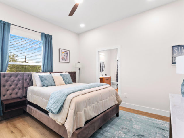 Bright modern bedroom with tufted upholstered bed, layered cream and teal bedding, teal curtains at a large window, ceiling fan, hardwood floors, patterned rug and an open doorway leading to an ensuite bathroom.