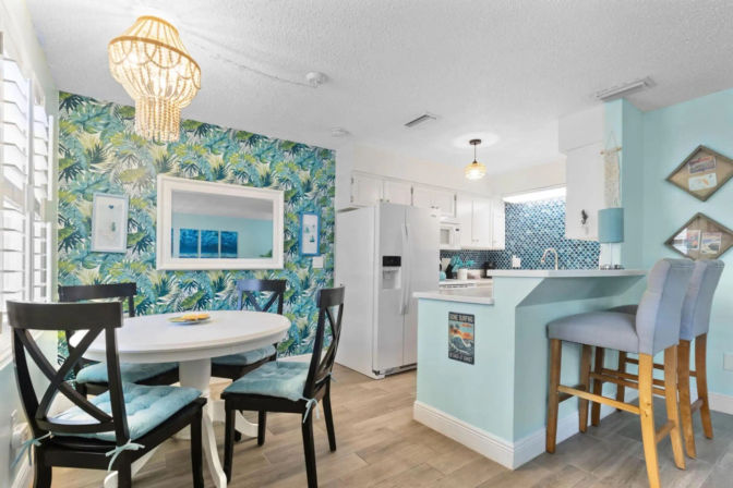 Bright beachy open-plan kitchen and dining area with tropical leaf wallpaper, round white table with black chairs, turquoise breakfast bar with upholstered stools, white appliances and pendant lights.