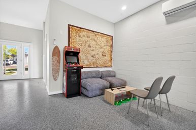Bright modern game lounge with a vintage arcade cabinet, striped gray modular sofa, carved wooden surfboard, large antique-style map on a white cinderblock wall, small wooden coffee table with green toy bins, two gray chairs, and glass doors letting in sunny outdoor light.