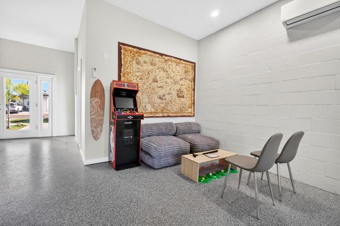 Bright modern game lounge with a vintage arcade cabinet, striped gray modular sofa, carved wooden surfboard, large antique-style map on a white cinderblock wall, small wooden coffee table with green toy bins, two gray chairs, and glass doors letting in sunny outdoor light.