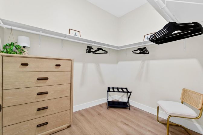 Bright modern walk-in closet with light wood dresser, floating shelf and black hangers, rattan chair, luggage rack and light hardwood floor.