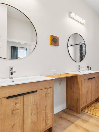 Contemporary bathroom double-sink vanity with round mirrors, chrome faucets, warm wood cabinets and a bright wall-mounted vanity light on neutral walls