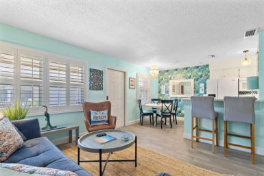 Bright, beachy open-plan living and dining area with mint walls and tropical wallpaper accent, blue sofa, leather armchair with 'To the Beach' pillow, round coffee table, shuttered windows, and barstools at a white kitchen peninsula.