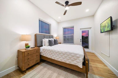Cozy modern bedroom with mid-century wooden bed and nightstand, white quilted bedding, ceiling fan, wall-mounted TV, hardwood floors and a glass door showing a pink-purple sunset.