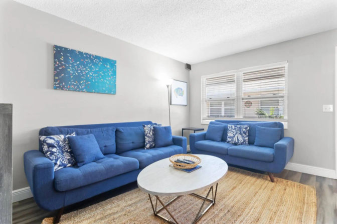Cozy modern living room with two vibrant blue sofas and patterned pillows, round white coffee table on a woven jute rug, large window with blinds and abstract blue wall art.