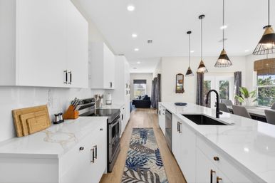 Bright modern open-concept kitchen with white cabinets, marble-look countertops, large island with black faucet, pendant lights, stainless-steel appliances, wood floors and a blue leaf runner.