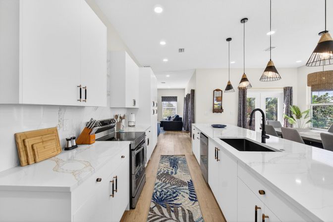 Bright modern open-concept kitchen with white cabinets, marble-look countertops, large island with black faucet, pendant lights, stainless-steel appliances, wood floors and a blue leaf runner.