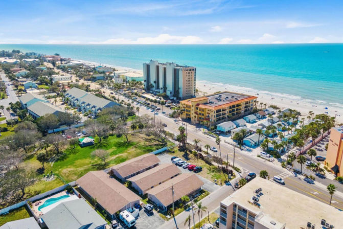 Aerial view of a sunny beach town with turquoise ocean, sandy shoreline, beachfront condos and colorful vacation cottages along a palm‑lined coastal road.