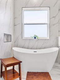 Bright modern bathroom with a white freestanding bathtub against marble walls, wooden bath stool and slatted mat, and a small potted plant on the frosted window sill.