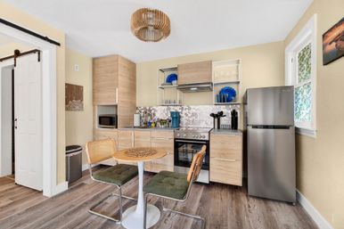 Cozy compact modern kitchen with light wood cabinets, stainless steel fridge and range, mosaic tile backsplash, round tulip table with two green-upholstered chairs and rattan accents, pendant light and wood-look flooring.