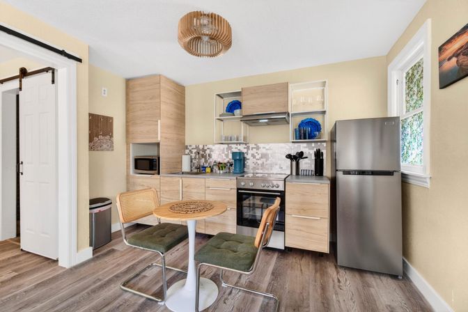 Cozy compact modern kitchen with light wood cabinets, stainless steel fridge and range, mosaic tile backsplash, round tulip table with two green-upholstered chairs and rattan accents, pendant light and wood-look flooring.