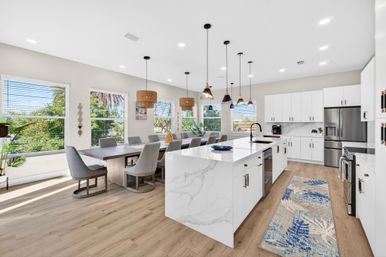 Sunlit open-plan modern kitchen and dining area with a white marble-look island and black faucet, stainless-steel appliances, white cabinets, multiple pendant lights, wood floors, and a long dining table with gray upholstered chairs beside large windows showing green foliage.