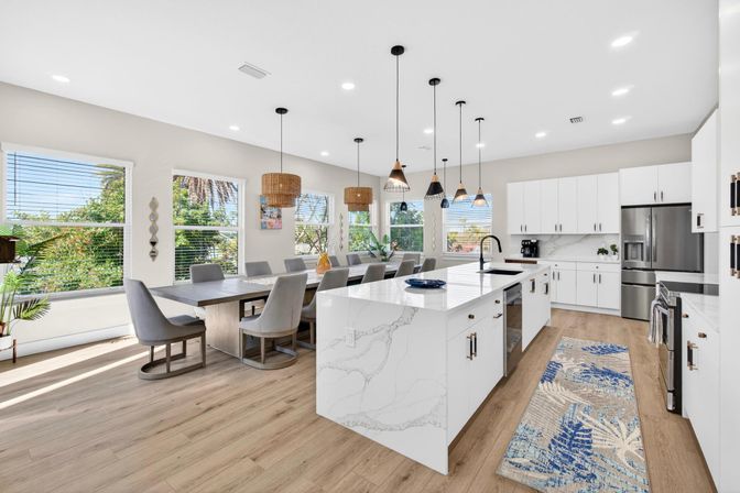 Sunlit open-plan modern kitchen and dining area with a white marble-look island and black faucet, stainless-steel appliances, white cabinets, multiple pendant lights, wood floors, and a long dining table with gray upholstered chairs beside large windows showing green foliage.