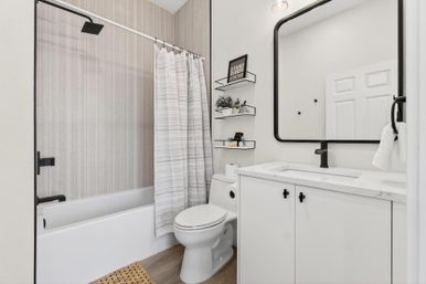 Bright modern bathroom with white bathtub and striped shower curtain, white vanity with marble countertop, black faucet and mirror frame, wall-mounted floating shelves with decor, toilet and wood-look flooring