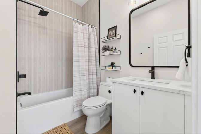 Bright modern bathroom with white bathtub and striped shower curtain, white vanity with marble countertop, black faucet and mirror frame, wall-mounted floating shelves with decor, toilet and wood-look flooring