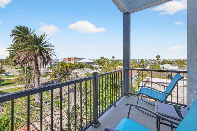 Sunny coastal balcony with bright blue lounge chairs and metal railing, overlooking palm trees, neighborhood rooftops and a distant waterway under a clear blue sky
