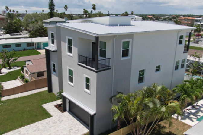 Modern three-story coastal home with a white metal roof, small balcony, ground-level garage, palm trees and paved driveway in a tropical neighborhood
