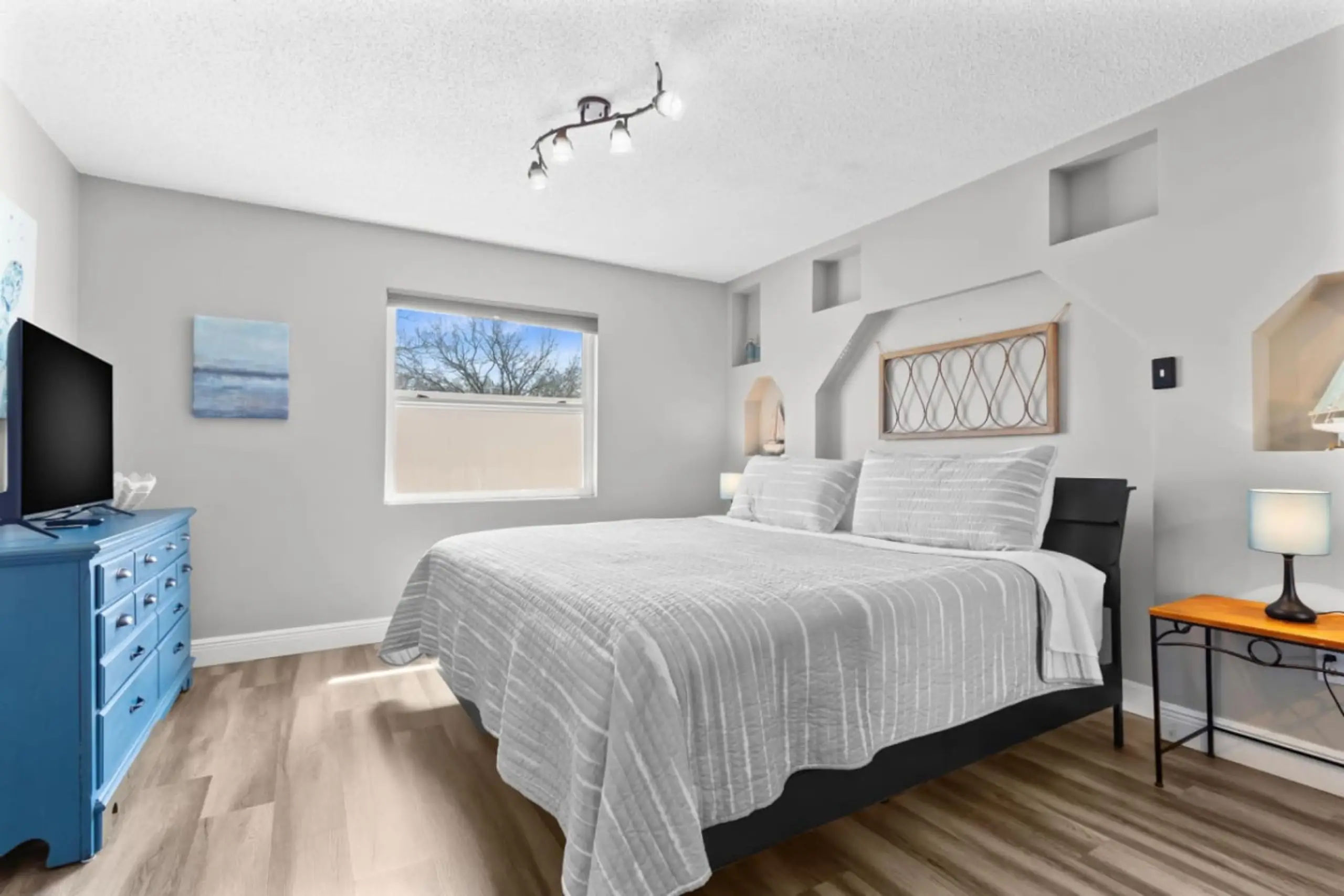 Sunlit modern bedroom with king bed and gray striped bedding, blue dresser, wood floors, window with tree view, wall niches and bedside lamps.