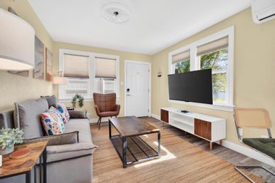Sunlit coastal living room with gray sofa and colorful pillows, leather accent chair, wood coffee table, wall-mounted TV, mid-century console, and palm-tree view through windows.