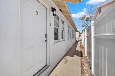 White exterior unit door marked 3 beside a row of windows and narrow sunlit walkway bordered by a white vinyl fence