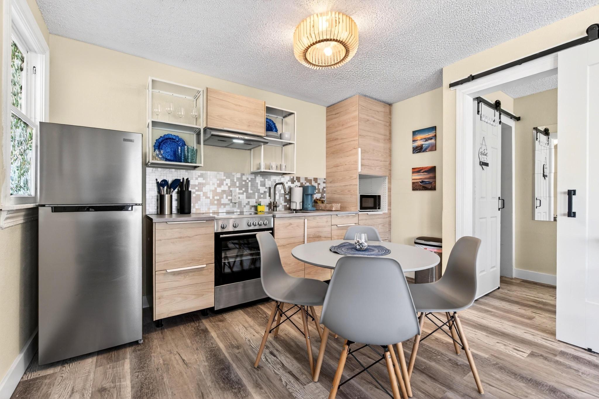 Bright modern apartment kitchen with light wood cabinets, stainless steel refrigerator and oven, mosaic tile backsplash, round dining table with four gray mid-century chairs, barn-style sliding door and wood-look flooring.