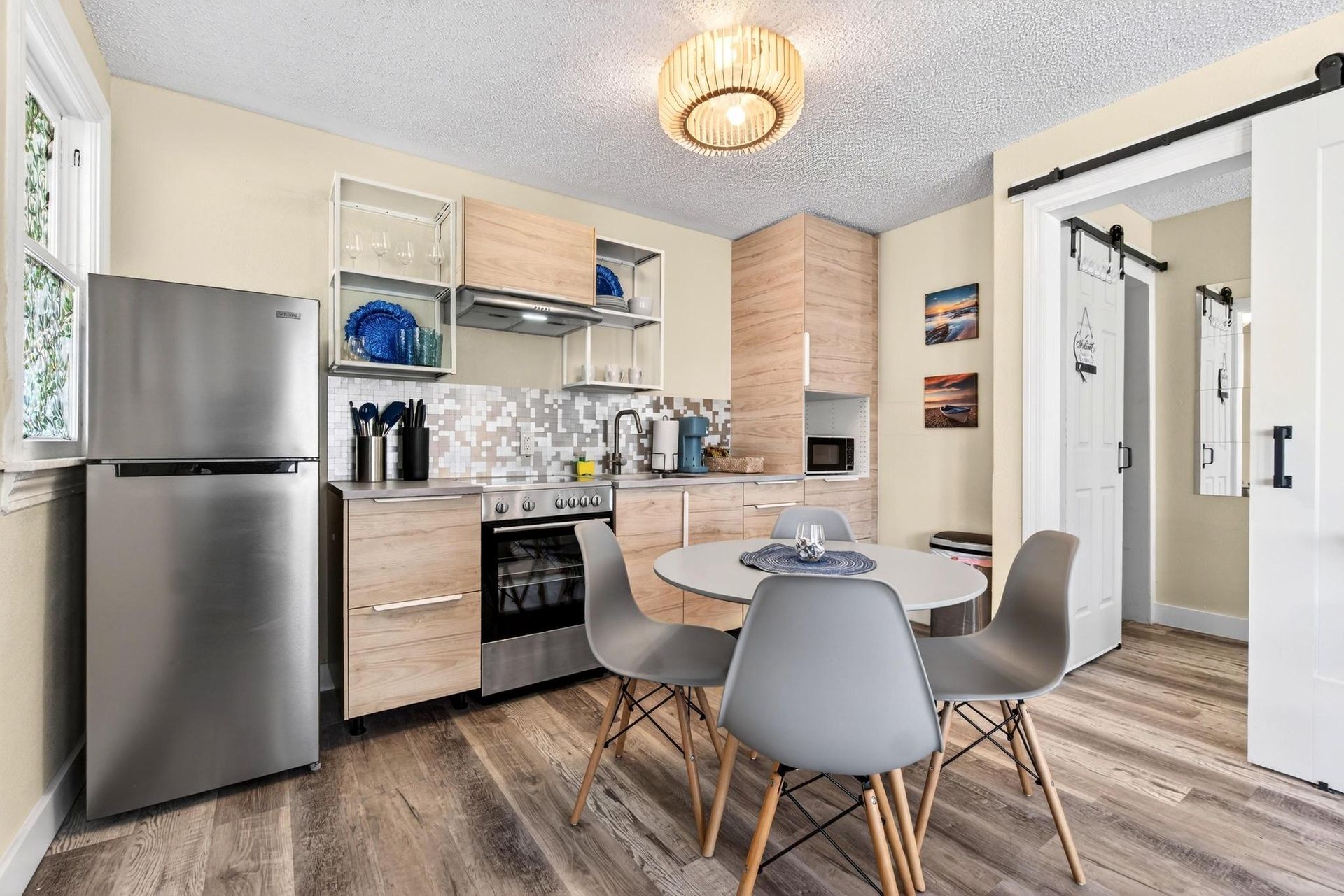 Bright modern apartment kitchen with light wood cabinets, stainless steel refrigerator and oven, mosaic tile backsplash, round dining table with four gray mid-century chairs, barn-style sliding door and wood-look flooring.