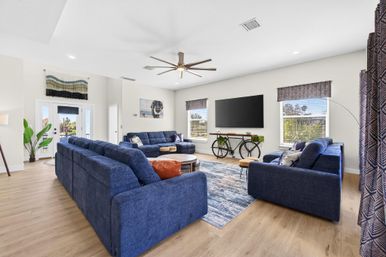 Spacious sunlit contemporary living room with navy sectional sofas and armchairs, light wood floors, patterned area rug, wall-mounted flat-screen TV above a bicycle-style console table, modern ceiling fan, potted plant by French doors, and large windows with valances.
