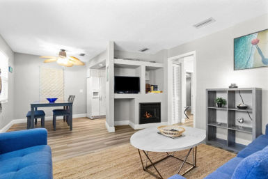Sunlit modern coastal-style apartment living room with blue sofas, round marble coffee table on woven rug, built-in TV above gas fireplace, small blue dining table, ceiling fan and light wood floors.