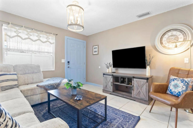 Cozy modern living room with a blue front door, light gray sectional, wooden coffee table on a blue patterned rug, wall-mounted TV on a rustic media console, and a leather accent chair under a large decorative wall clock.