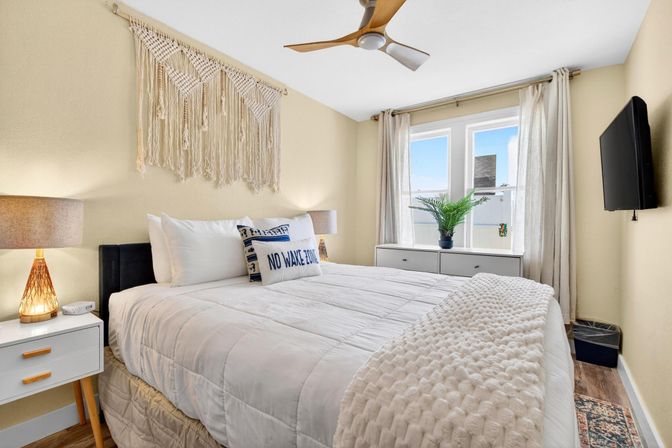 Sunlit cozy coastal-style bedroom with white quilted bed, "NO WAKE ZONE" accent pillow, macramé wall hanging, wooden ceiling fan, bedside lamps on mid-century nightstands, sheer-curtained window with potted plant and wall-mounted TV.