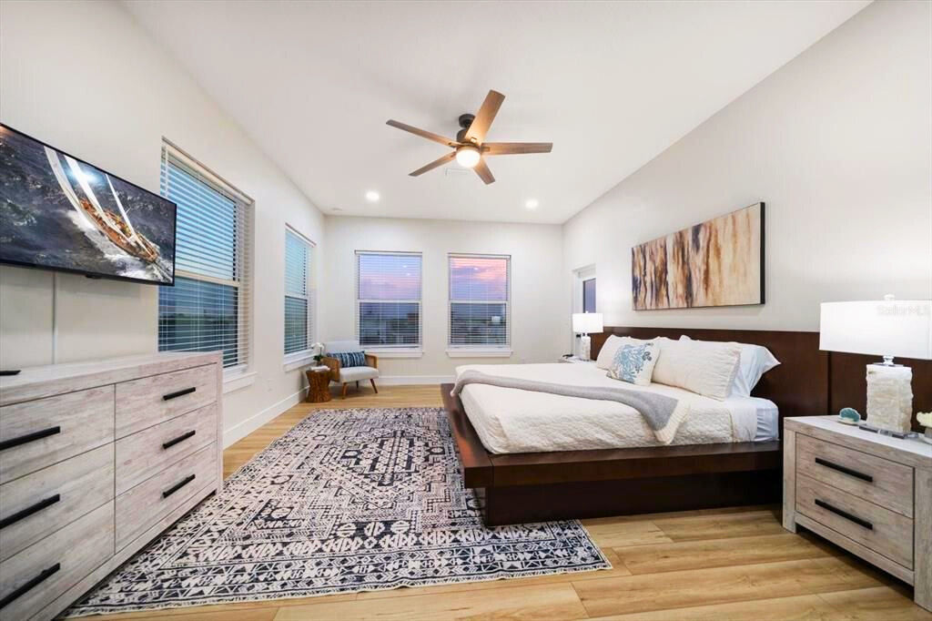 Bright contemporary master bedroom with low wood platform bed, patterned area rug, ceiling fan, dresser with TV and three windows showing a sunset view