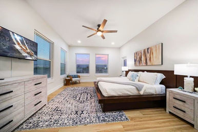 Bright contemporary master bedroom with low wood platform bed, patterned area rug, ceiling fan, dresser with TV and three windows showing a sunset view